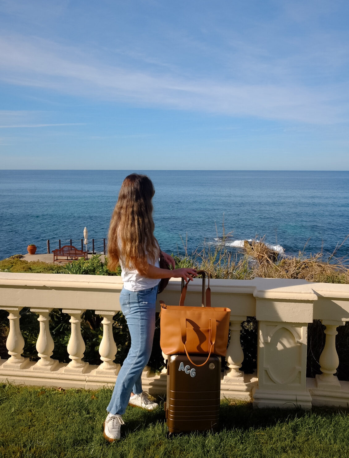 Alexandra Grant posing with Monos hybrid carry-on luggage and metro tote in scenic ocean setting in Sardinia, Italy.