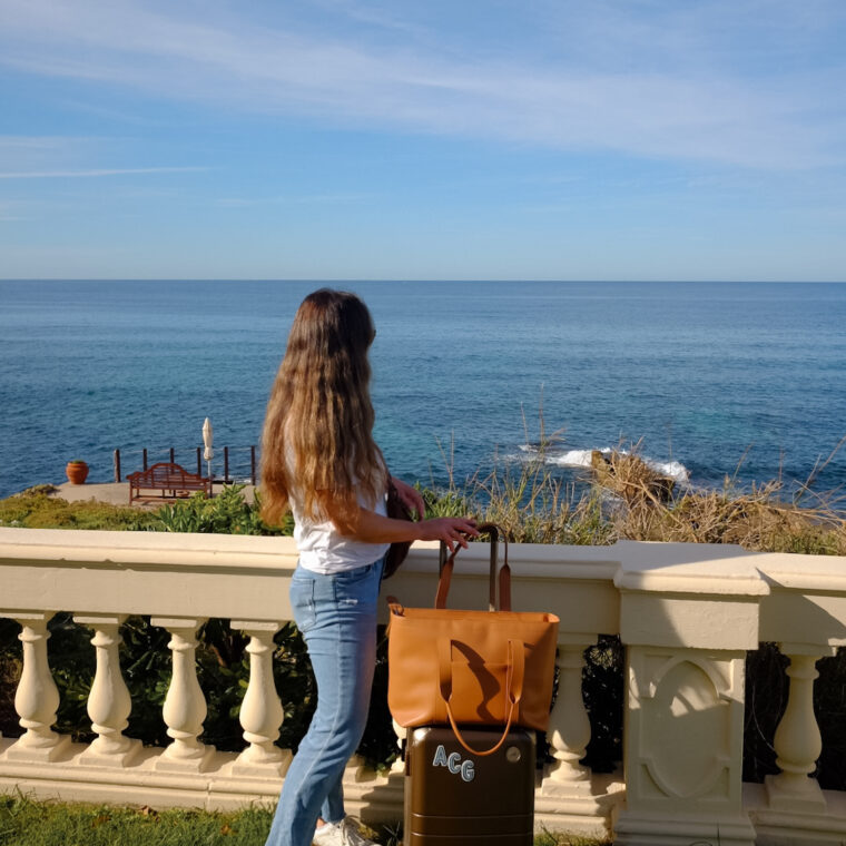 Alexandra Grant posing with Monos hybrid carry-on luggage and metro tote in scenic ocean setting in Sardinia, Italy.