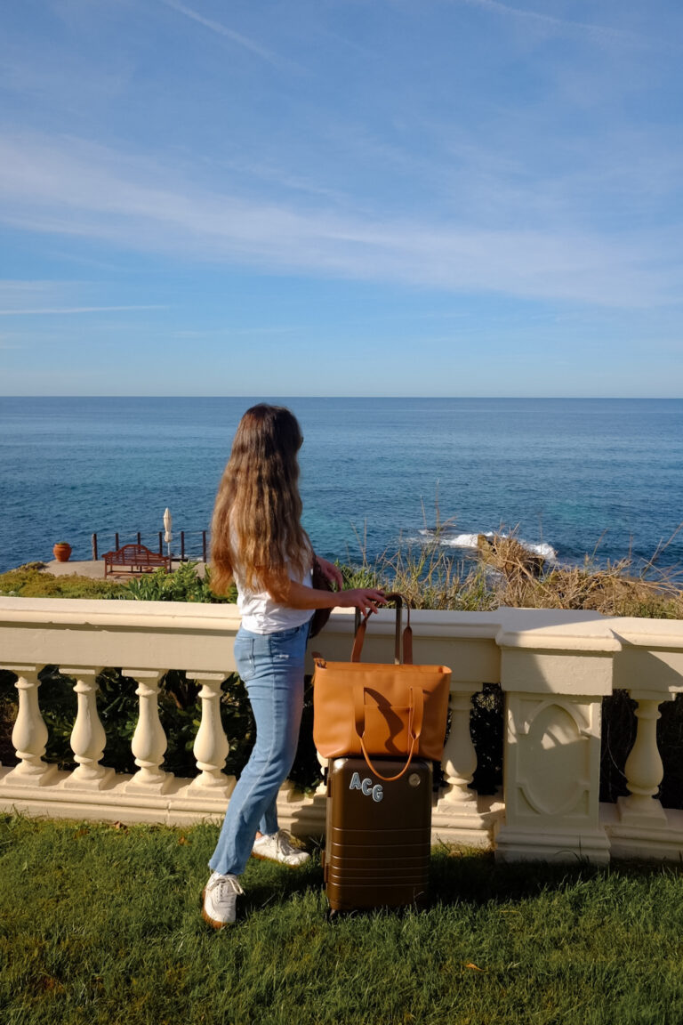 Alexandra Grant posing with Monos hybrid carry-on luggage and metro tote in scenic ocean setting in Sardinia, Italy.