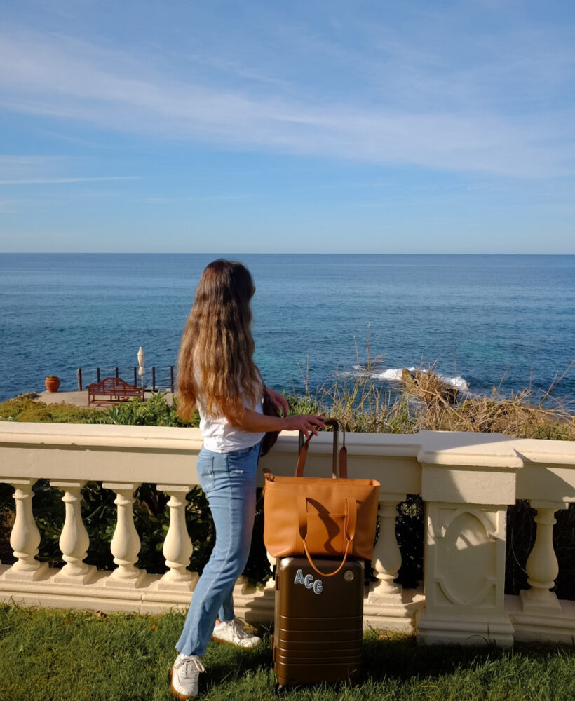 Alexandra Grant posing with Monos hybrid carry-on luggage and metro tote in scenic ocean setting in Sardinia, Italy.