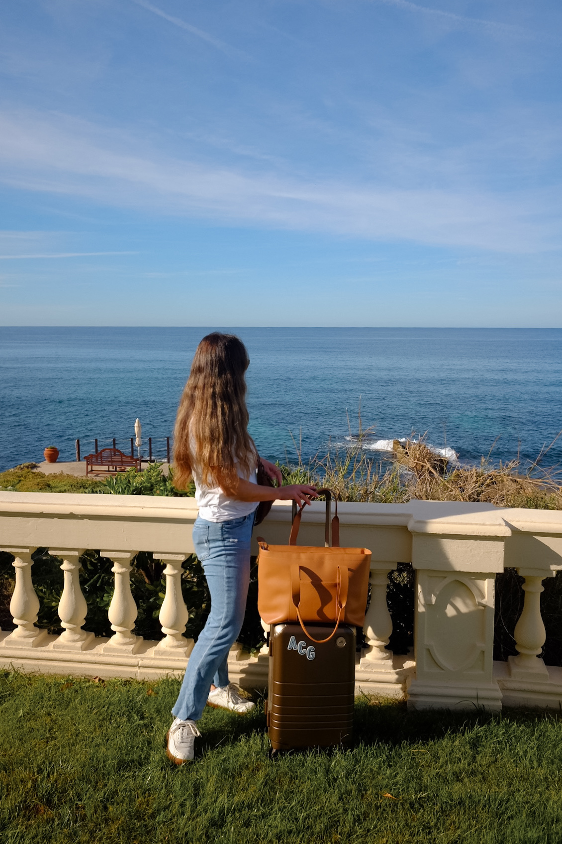 Alexandra Grant posing with Monos hybrid carry-on luggage and metro tote in scenic ocean setting in Sardinia, Italy.